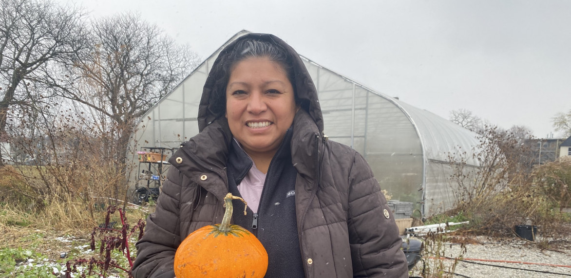 A woman stands in a garden wearing a winter coat and holding an orange pumpkin. 